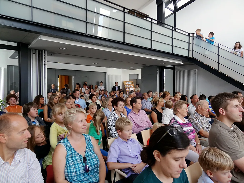 Voll besetzte Aula mit Familien, auf der Empore weitere Zuschauer.